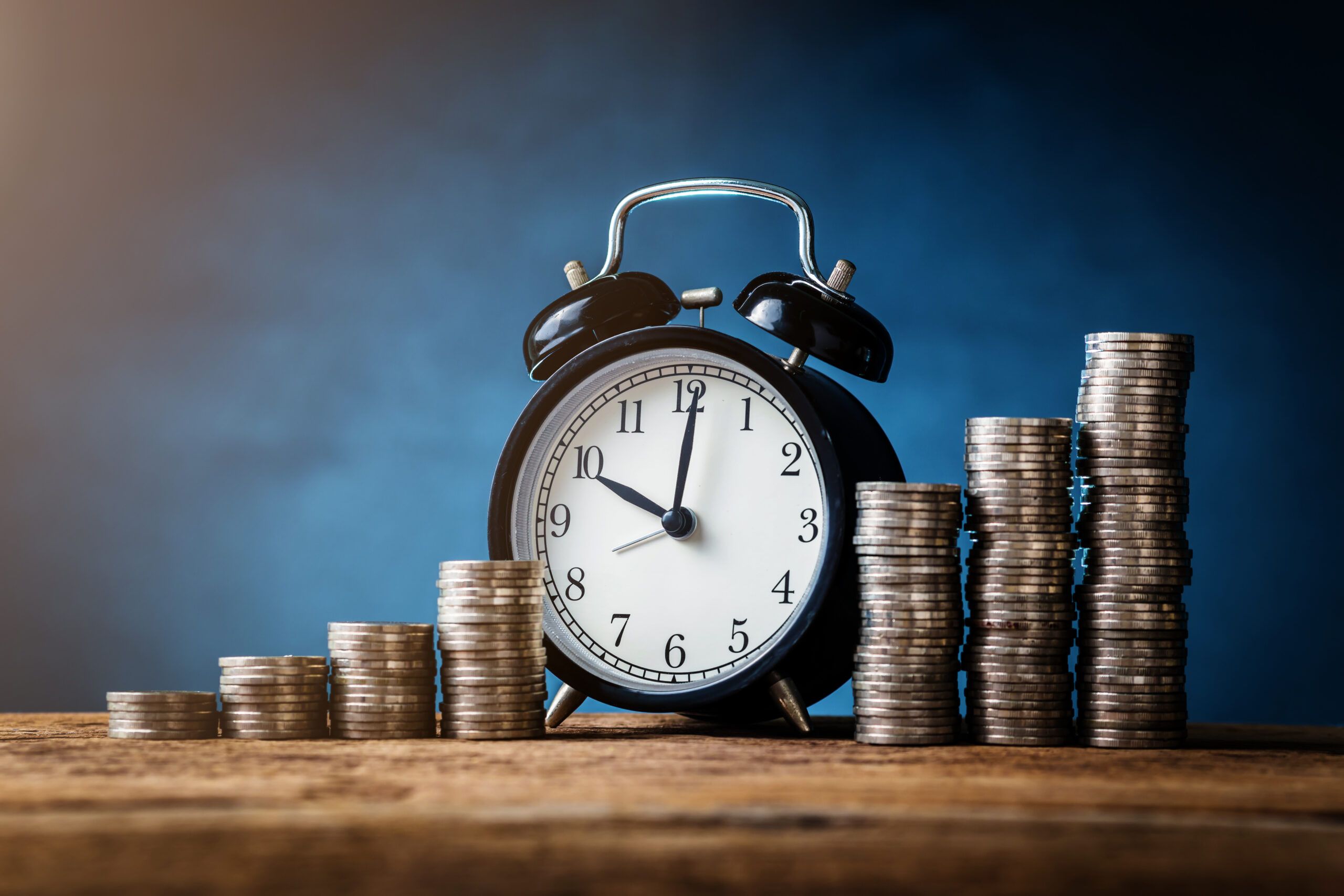 A stock photograph showing an alarm clock amidst a pile of money, which has been arranged in ascending increments.