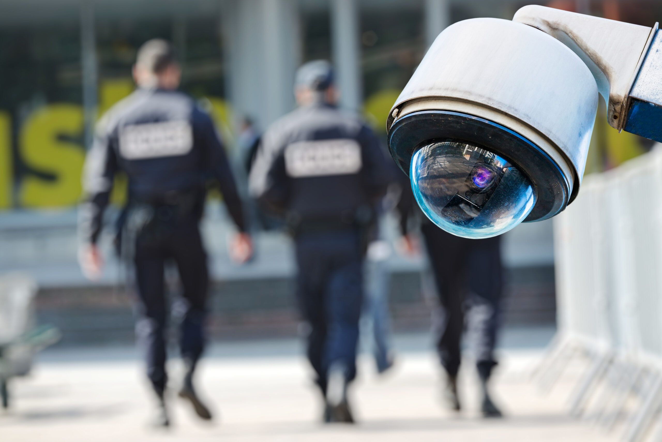 Three security officers patrol in the blurred background. In the foreground, a fisheye lens CCTV unit watches the camer.